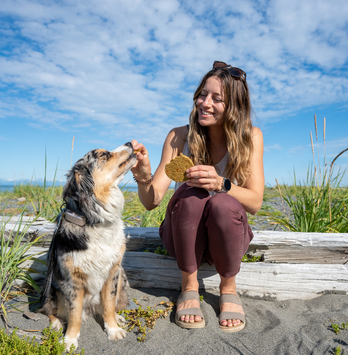 Woman sitting on a log with a dog on a beach, promoting Artisan Raw food.