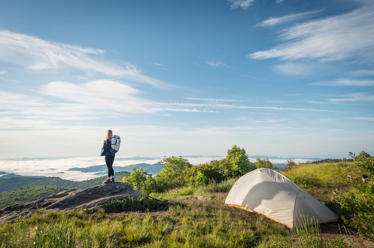 A person stands on a rock near a tent, overlooking a mountainous landscape with clouds below and a blue sky above.